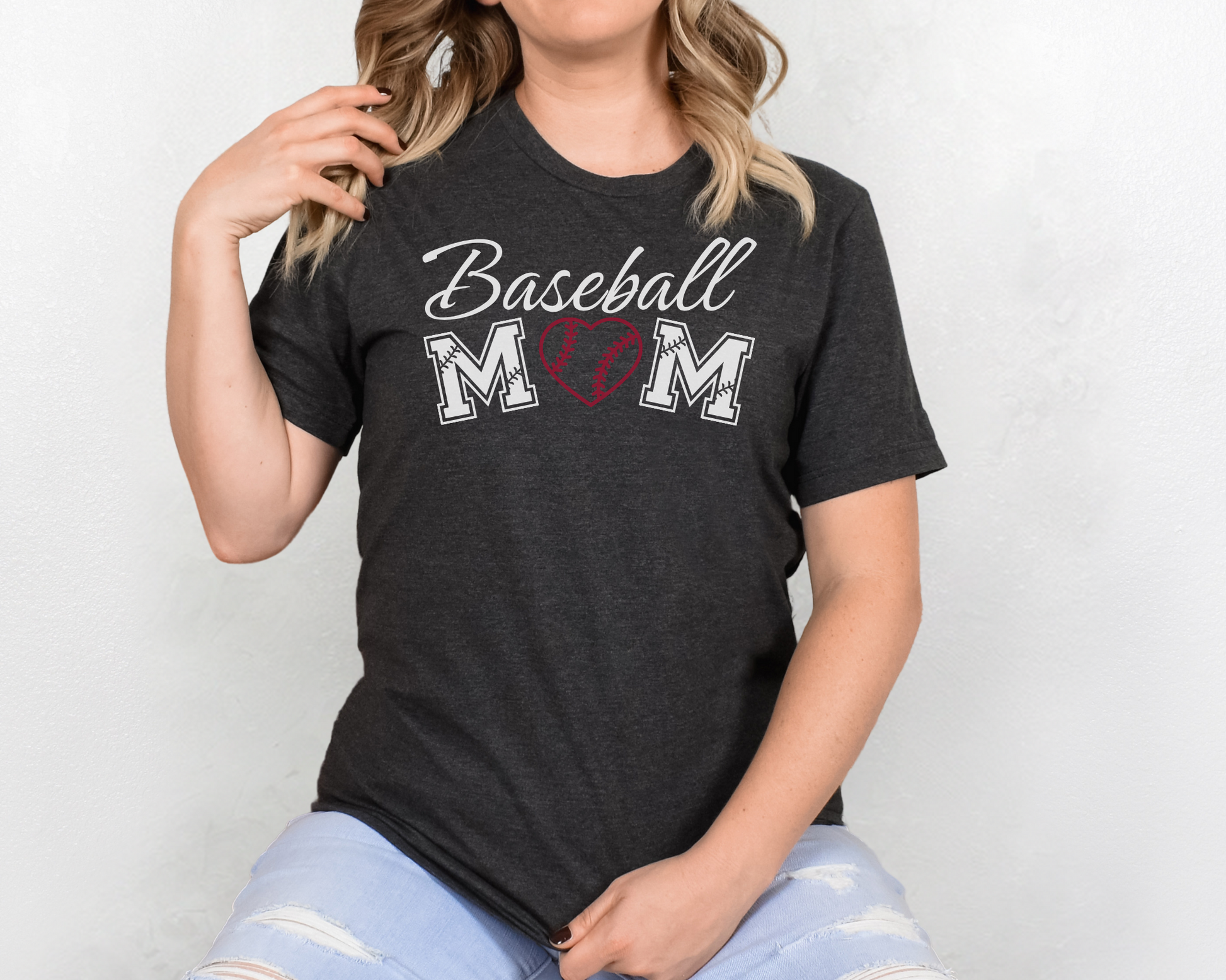 A model wearing a dark gray t-shirt with a 'Baseball Mom' design. The design features white cursive font for "baseball" and white baseball font for "mom". A red baseball heart replaces the "O" in "mom".