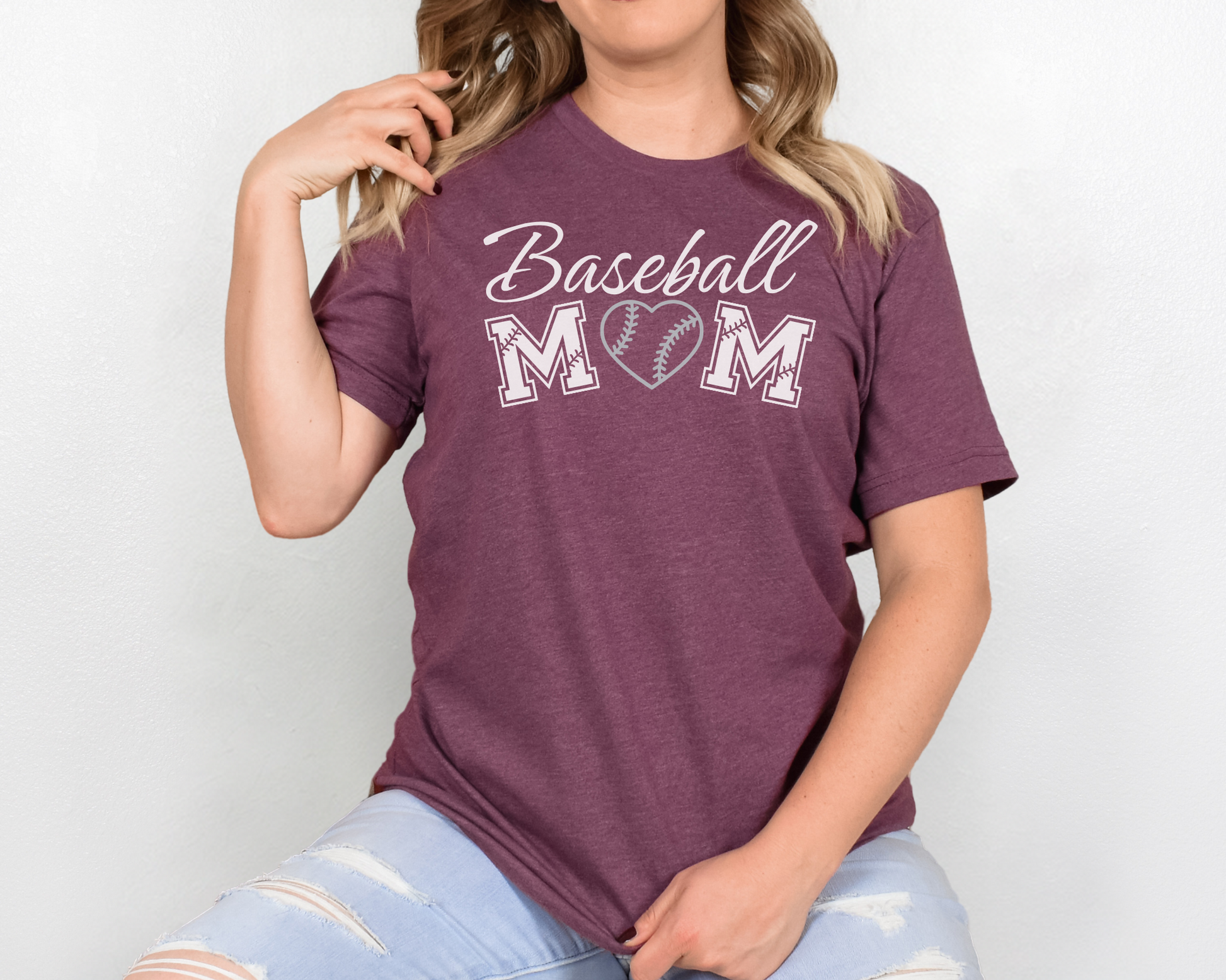 A model wearing a maroon t-shirt with a 'Baseball Mom' design. The design features white cursive font for "baseball" and white baseball font for "mom". A silver baseball heart replaces the "O" in "mom".