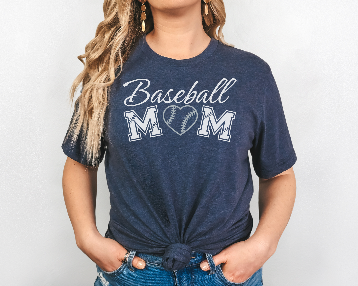 A model wearing a navy t-shirt with a 'Baseball Mom' design. The design features white cursive font for "baseball" and white baseball font for "mom". A silver baseball heart replaces the "O" in "mom".