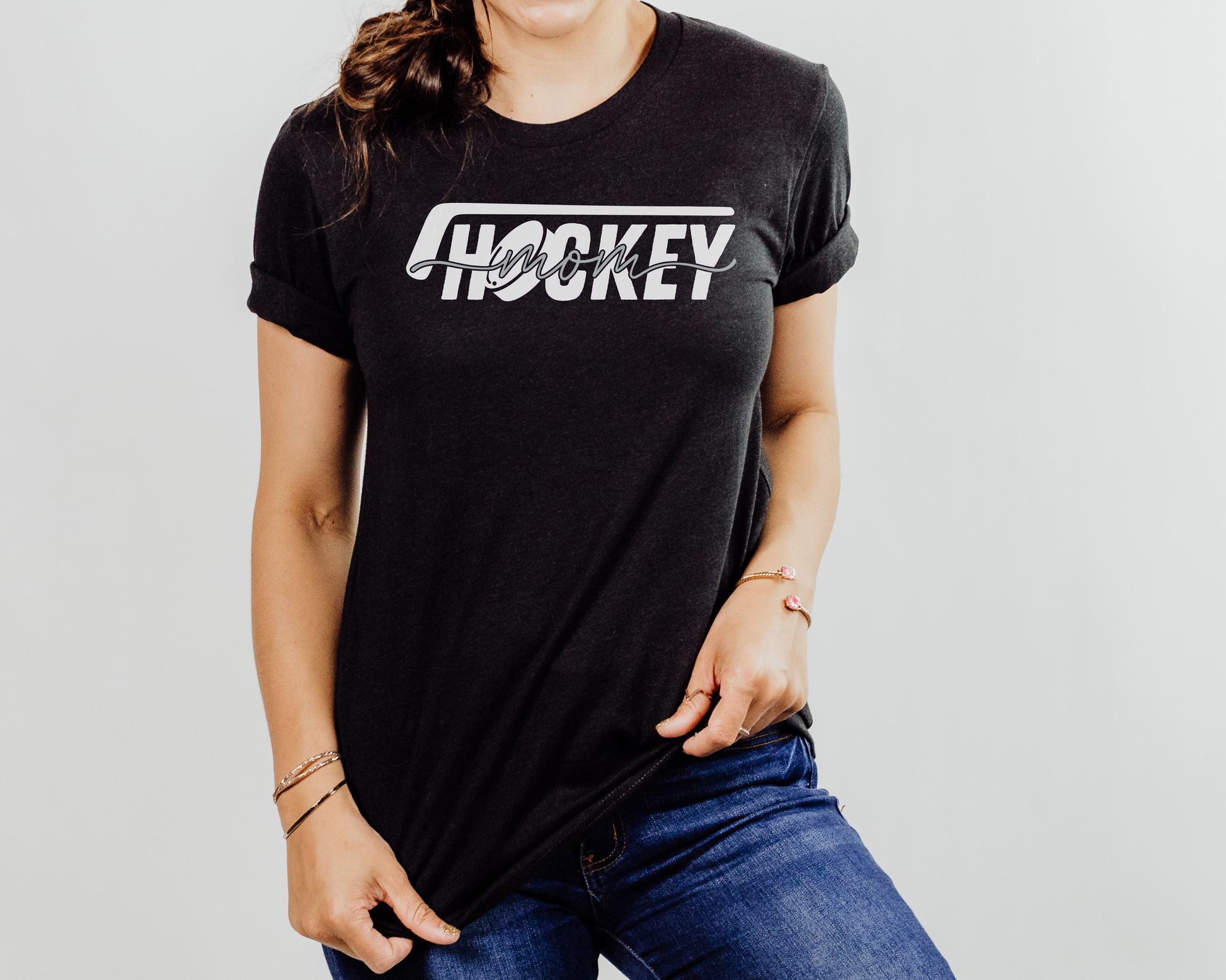 A model wearing a black t-shirt with a 'Hockey Mom' design. The design features a white hockey stick above the word 'hockey', with a puck representing the 'O' in hockey. The word 'Mom' overlays across the word 'hockey'.