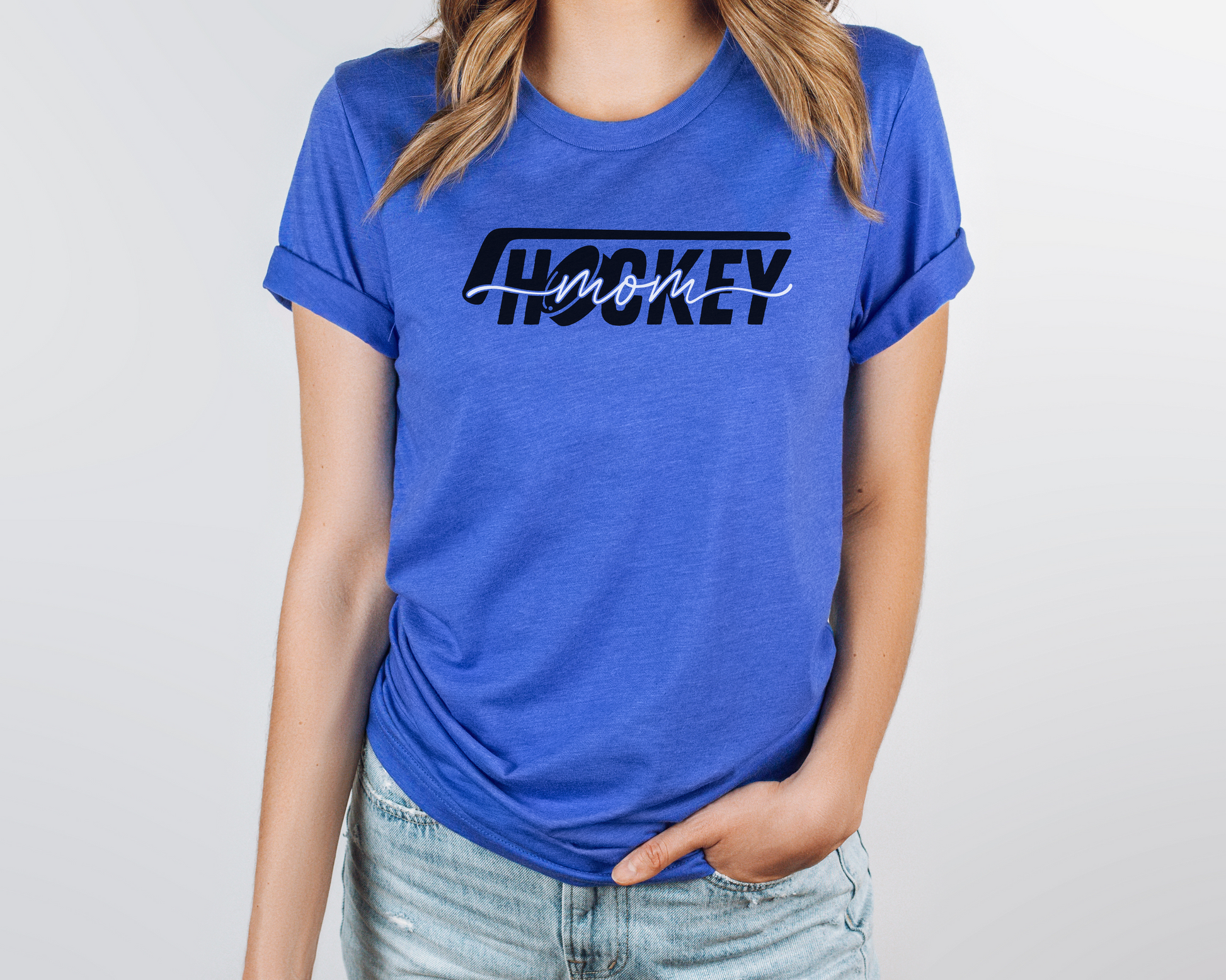 A model wearing a heather blue t-shirt with a 'Hockey Mom' design. The design features a black hockey stick above the word 'hockey', with a puck representing the 'O' in hockey. The word 'Mom' overlays across the word 'hockey'.