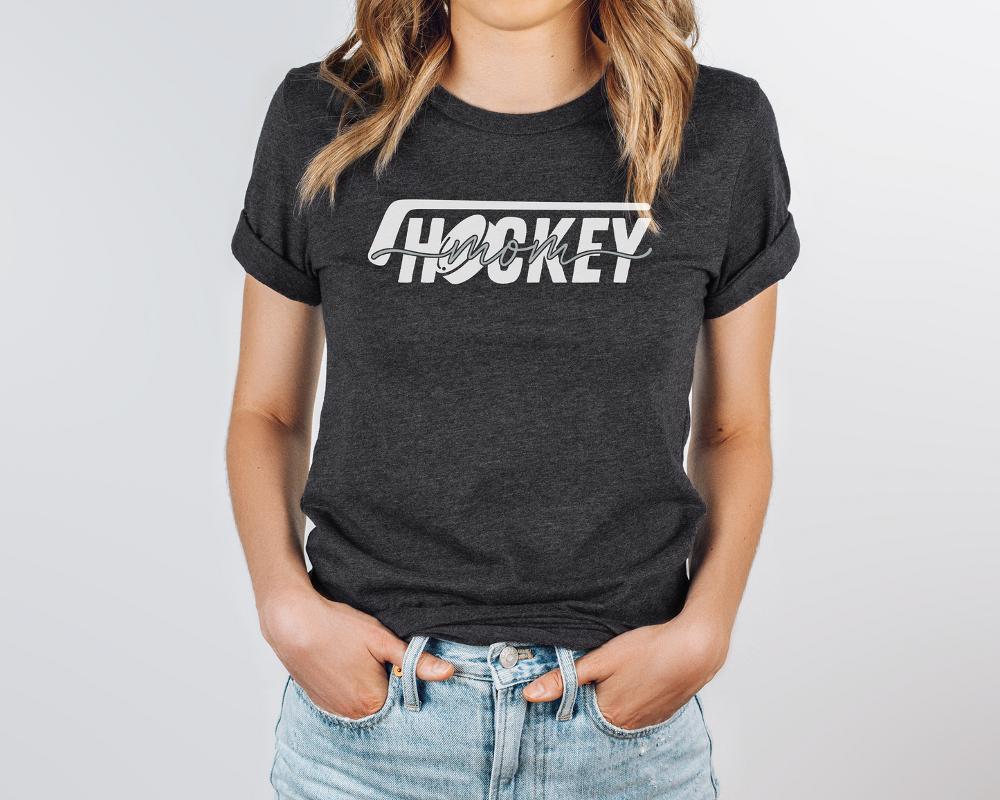 A model wearing a dark gray heather t-shirt with a 'Hockey Mom' design. The design features a white hockey stick above the word 'hockey', with a puck representing the 'O' in hockey. The word 'Mom' overlays across the word 'hockey'.