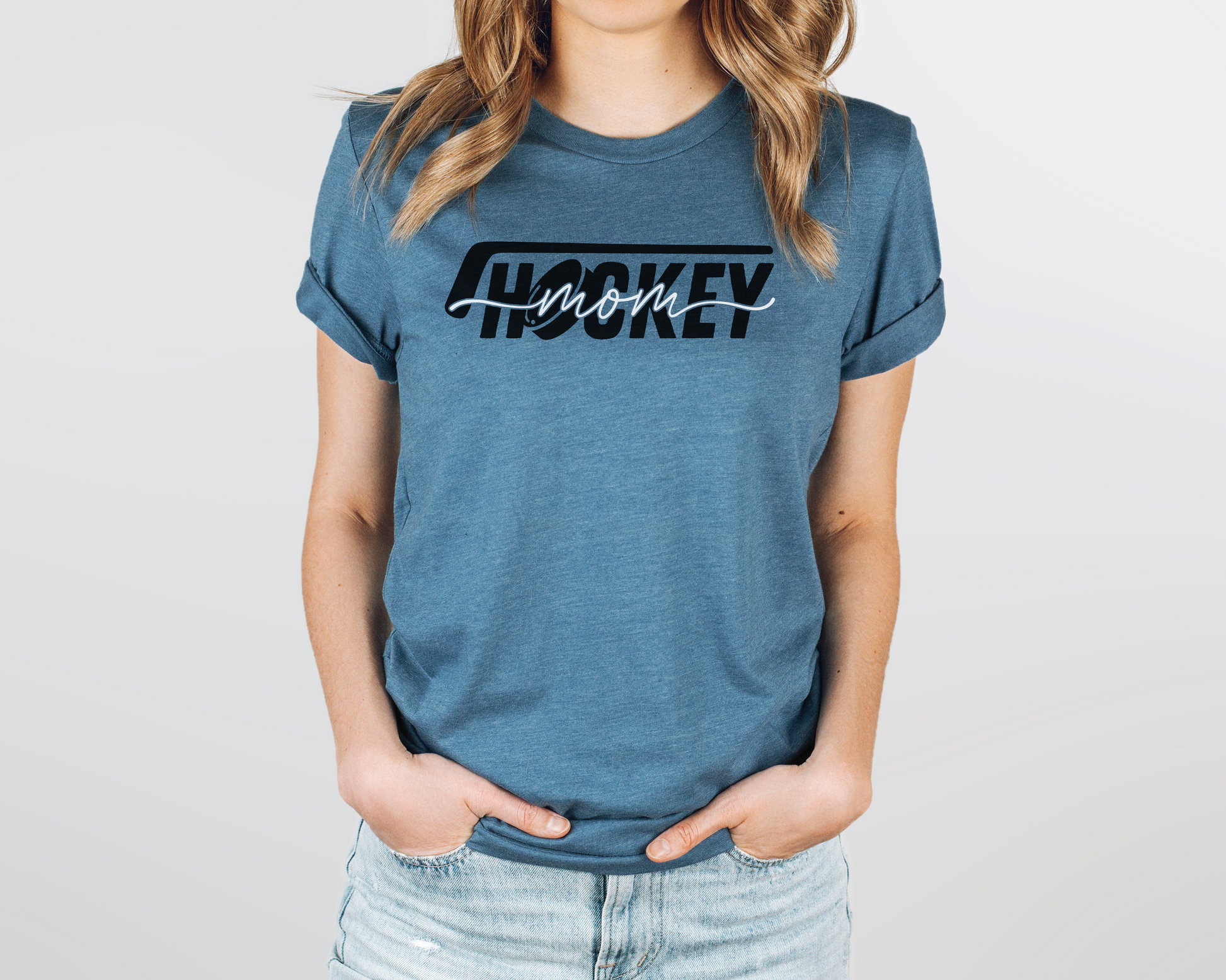 A model wearing a heather dark teal t-shirt with a 'Hockey Mom' design. The design features a black hockey stick above the word 'hockey', with a puck representing the 'O' in hockey. The word 'Mom' overlays across the word 'hockey'.