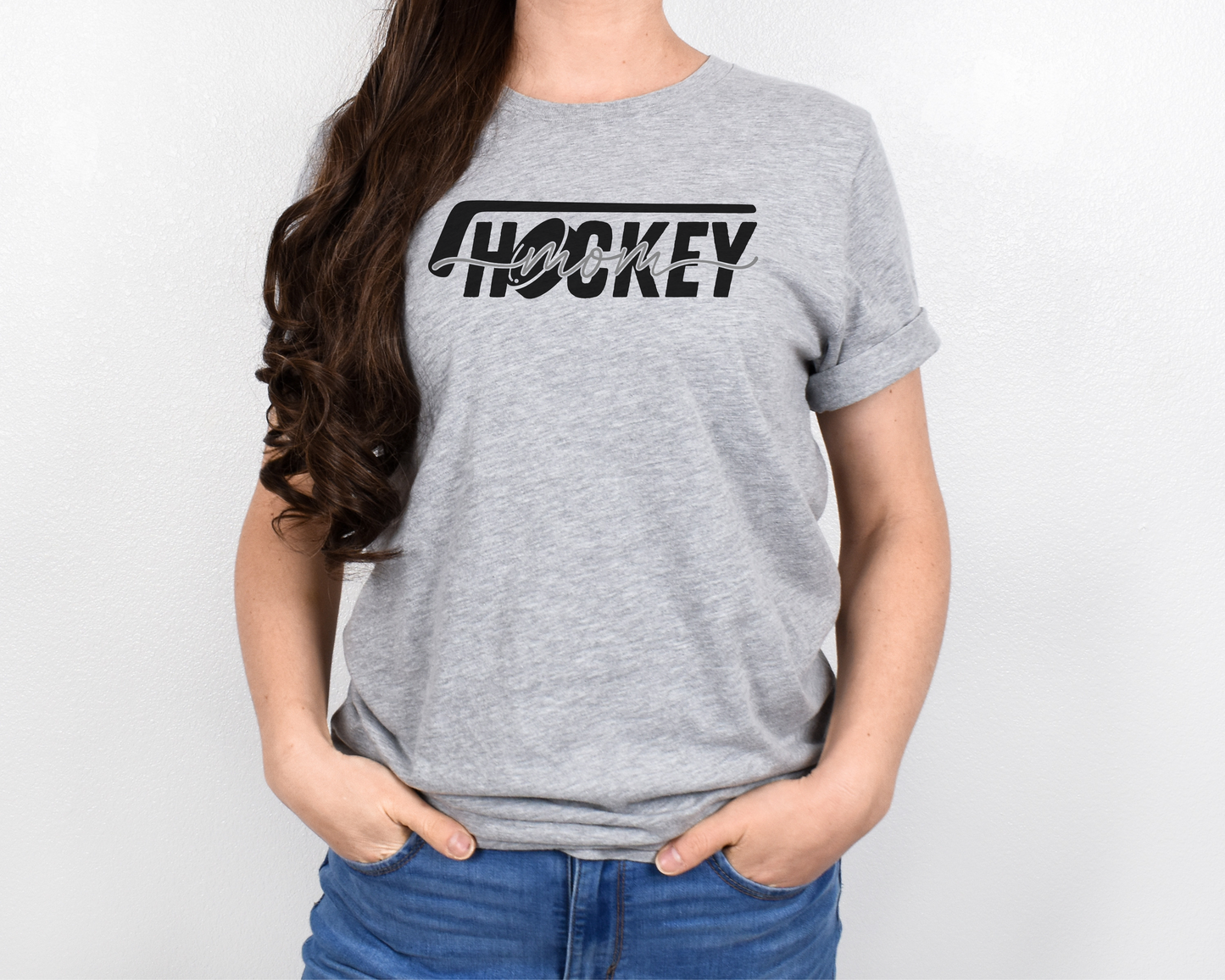 A model wearing a heather light gray t-shirt with a 'Hockey Mom' design. The design features a black hockey stick above the word 'hockey', with a puck representing the 'O' in hockey. The word 'Mom' overlays across the word 'hockey'.