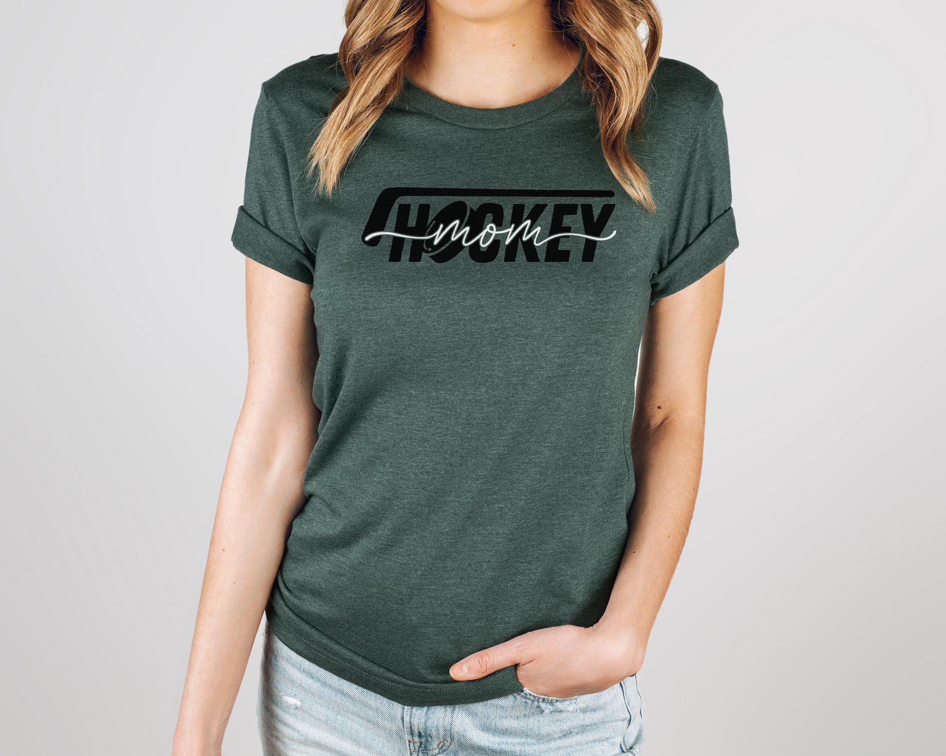 A model wearing a green heather t-shirt with a 'Hockey Mom' design. The design features a black hockey stick above the word 'hockey', with a puck representing the 'O' in hockey. The word 'Mom' overlays across the word 'hockey'.