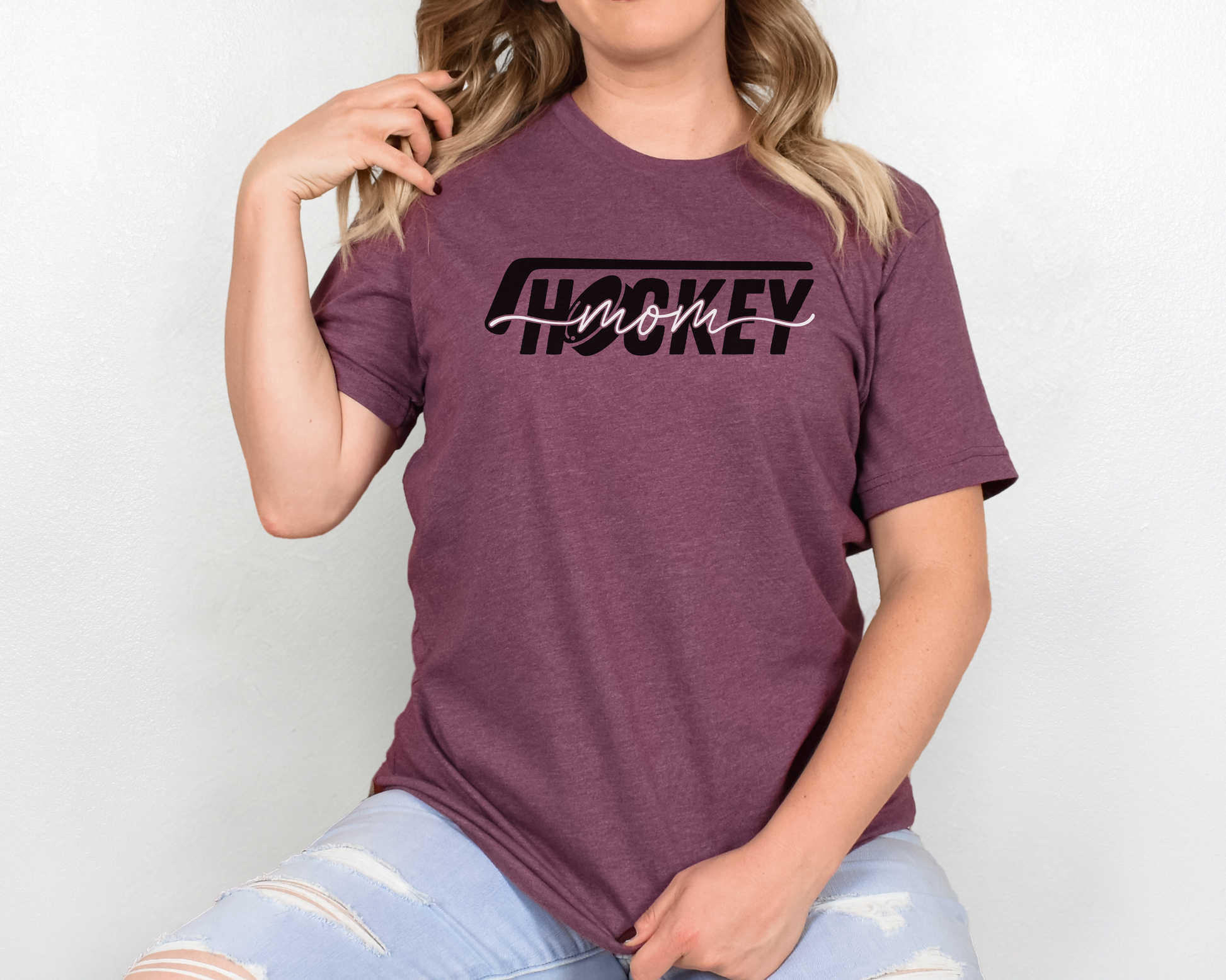 A model wearing a heather maroon t-shirt with a 'Hockey Mom' design. The design features a black hockey stick above the word 'hockey', with a puck representing the 'O' in hockey. The word 'Mom' overlays across the word 'hockey'.