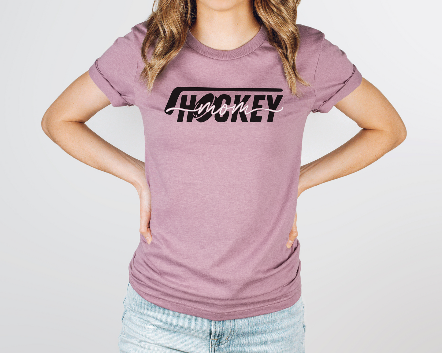 A model wearing a pink heather t-shirt with a 'Hockey Mom' design. The design features a black hockey stick above the word 'hockey', with a puck representing the 'O' in hockey. The word 'Mom' overlays across the word 'hockey'.