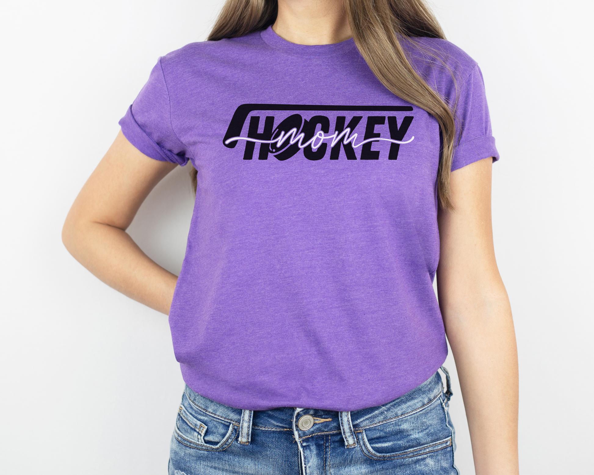 A model wearing a heather purple t-shirt with a 'Hockey Mom' design. The design features a black hockey stick above the word 'hockey', with a puck representing the 'O' in hockey. The word 'Mom' overlays across the word 'hockey'.