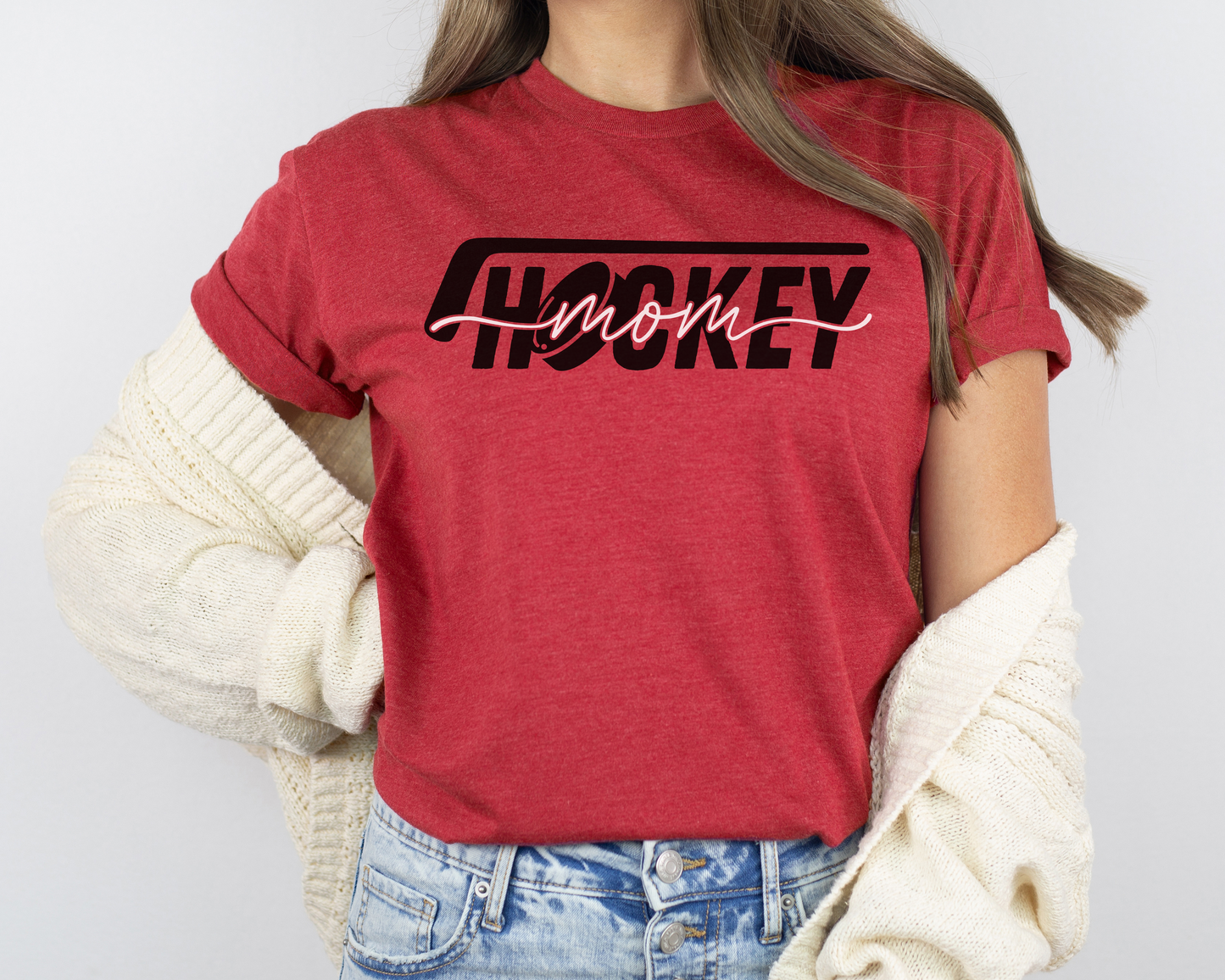 A model wearing a red heather t-shirt with a 'Hockey Mom' design. The design features a black hockey stick above the word 'hockey', with a puck representing the 'O' in hockey. The word 'Mom' overlays across the word 'hockey'.