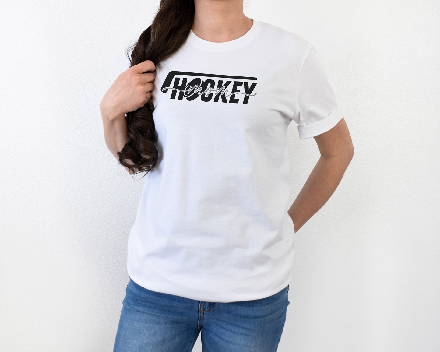 A model wearing a white t-shirt with a 'Hockey Mom' design. The design features a black hockey stick above the word 'hockey', with a puck representing the 'O' in hockey. The word 'Mom' overlays across the word 'hockey'.