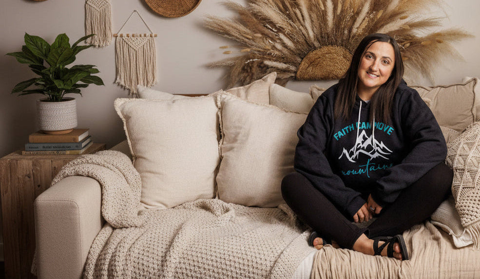 A woman sits cross-legged on a beige bed with pillows and blankets, wearing a black hoodie with a blue graphic that reads 'Faith Can Move Mountains'. The background features a large dried pampas grass wall decor piece.