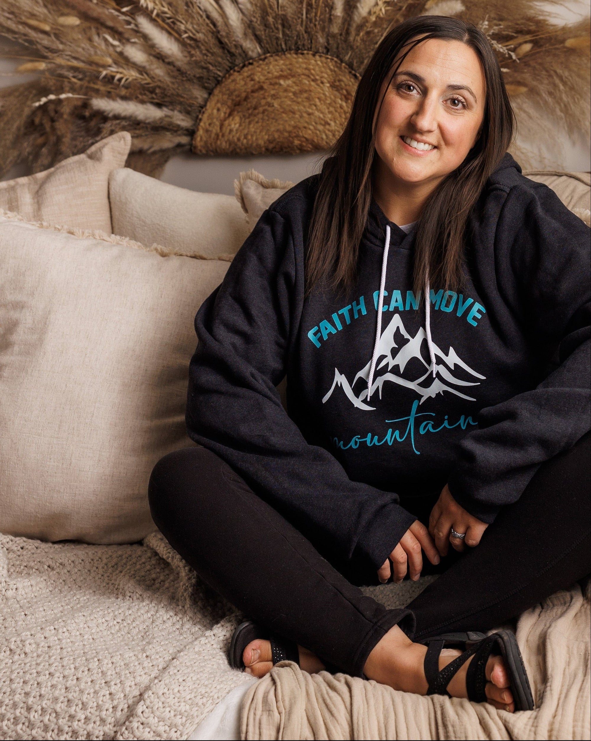 A woman sits cross-legged on a beige bed with pillows and blankets, wearing a black hoodie with a blue graphic that reads 'faith can move mountains'. The background features a large dried pampas grass wall decor piece.