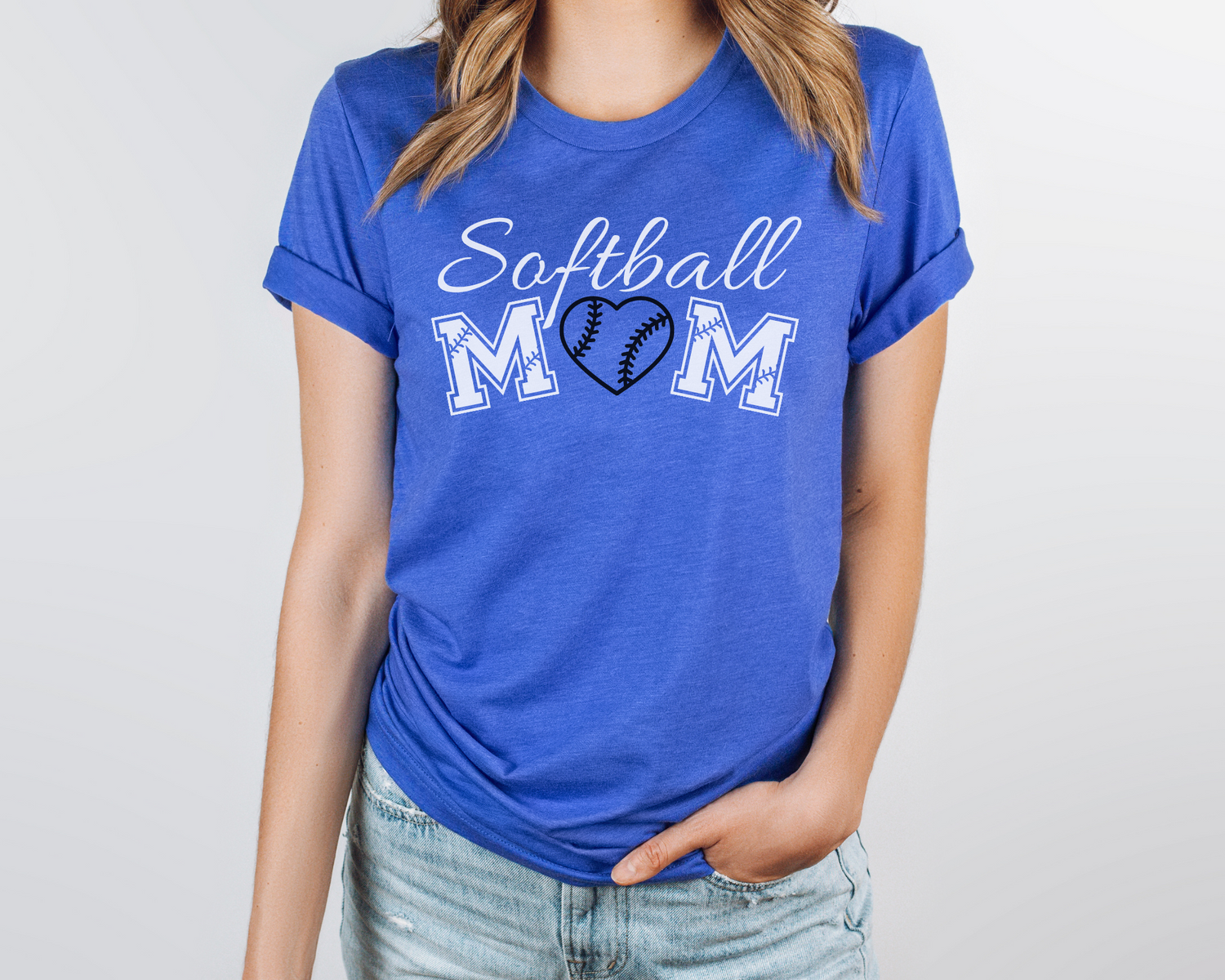 A model wearing a blue t-shirt with a 'Softball Mom' design. The design features white cursive font for "softball" and white softball font for "mom". A black softball heart replaces the "O" in "mom".